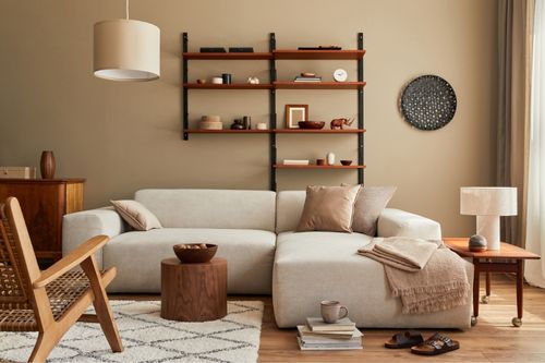 A living room designed with earth tones. The rattan chair and side tables are a wood-toned brown, the walls are painted with a warm latte color, the pillows and couch range from off-white to clay, and the hardwood floor is a light-stained wood. Black accents round out the room which has a calm and cozy ambience.