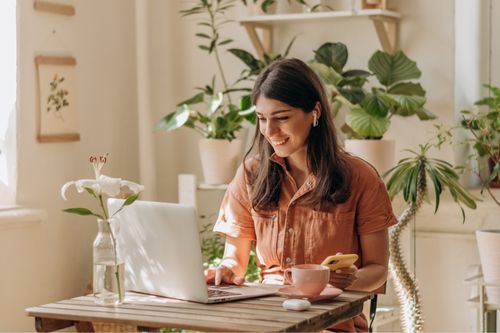 A young woman works from home on her laptop surrounded by house plants. The walls are off white, and with white plaster planters and natural wood accents, the green color of the plants pops against the clean backdrop.