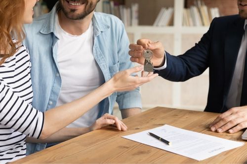 In a small office, a real estate agent hands the keys to a new home to their clients. The real estate contract is on the table in front of them.