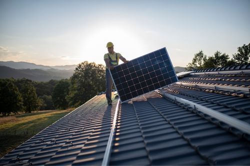 A worker installs a solar panel on the rooftop of a sustainable home as the sun sets behind him.