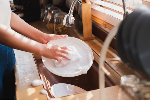 A woman practices sustainable habits by washing a plate in her kitchen sink. The sponge is full of soap and the water is off while she scrubs the plate.