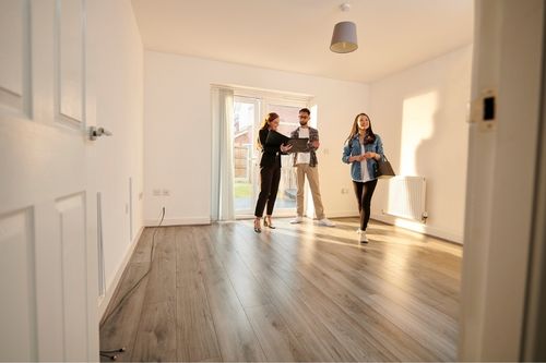 A man and a woman walk through a house for rent with the landlord. The landlord shows paperwork while they explore the living room. It is a new construction home with white walls and hardwood flooring.