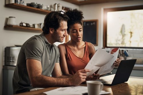 A young interracial heterosexual couple sits at their dining room table in the open concept kitchen of their new home reviewing their home warranty policy printed out with their laptop open.