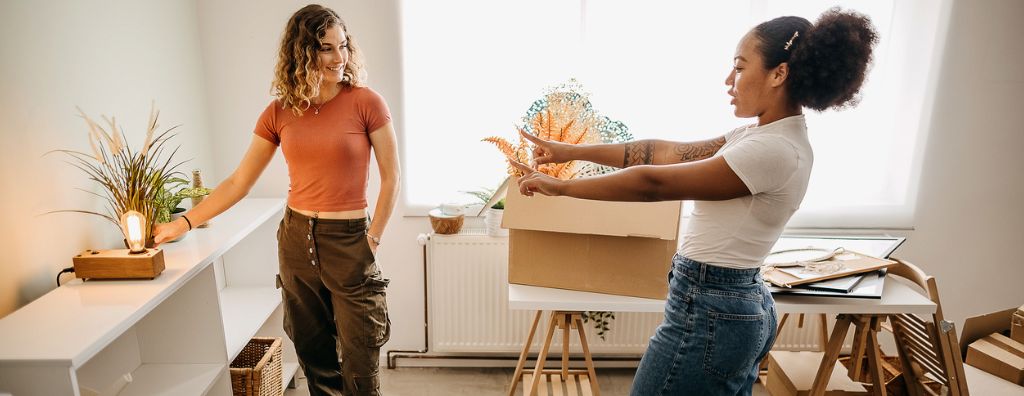 Two young women unpack boxes and arrange the living room as they move into a house they are renting. There are modern decorations including an exposed lightbulb lamp, bookshelf, dining room table, and a few small house plants. Two young women unpack boxes and arrange the living room as they move into a house they are renting. There are modern decorations including an exposed lightbulb lamp, bookshelf, dining room table, and a few small house plants.