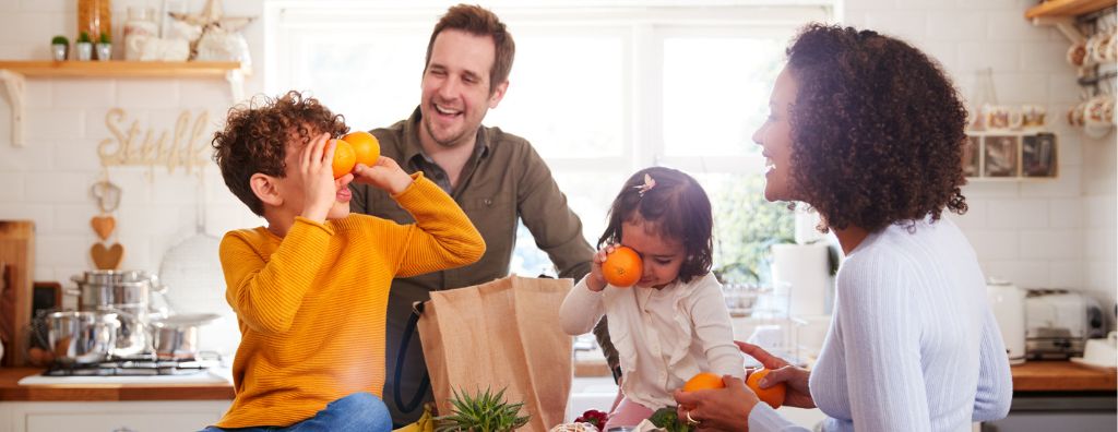 A family returns home after going grocery shopping with reusable bags. The son and daughter play with fresh oranges purchased from a natural market. There are houseplants on the kitchen island and appliances and utensils on the counters.