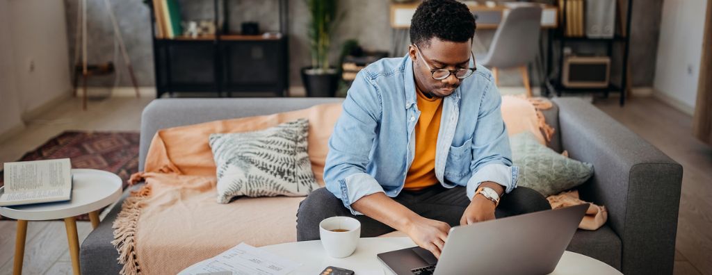 A young black man reads his home warranty on his laptop while he sits in his living room. The coffee table has notes and his phone, as well as a cup of coffee. The living room is decorated with modern furniture and house plants.