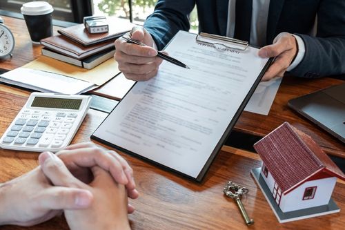 A close up shot of a real estate agent presenting a pre-approval mortgage agreement to their client. There is a calculator, a key, and a small model of a house on the real estate agent’s desk.