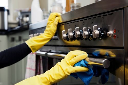 A closeup shot of a person’s hands. Wearing yellow rubber gloves, they clean the face of an electric stove and oven range.