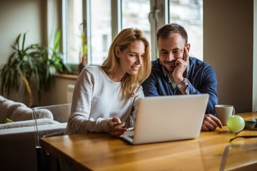 A young heterosexual Caucasian couple view real estate listings on their laptop at the dining room table. The home is decorated with modern furniture and house plants.