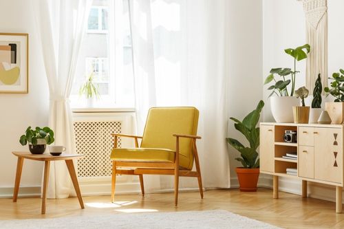 A mid-century modern living room with an open credenza containing cameras and books, a comfortable yellow chair with wooden peg legs, a small minimalist table, and several house plants. The colors of the furniture and plants pop against the white walls and white sheer curtains.