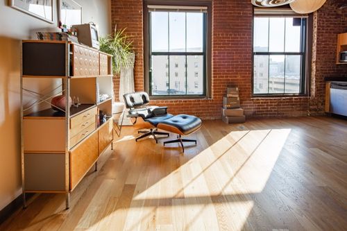 A leather Eames chair in a modern brick loft apartment with hardwood floors, an open kitchen/dining room area, and a large bookshelf decorated with accent items and house plants.