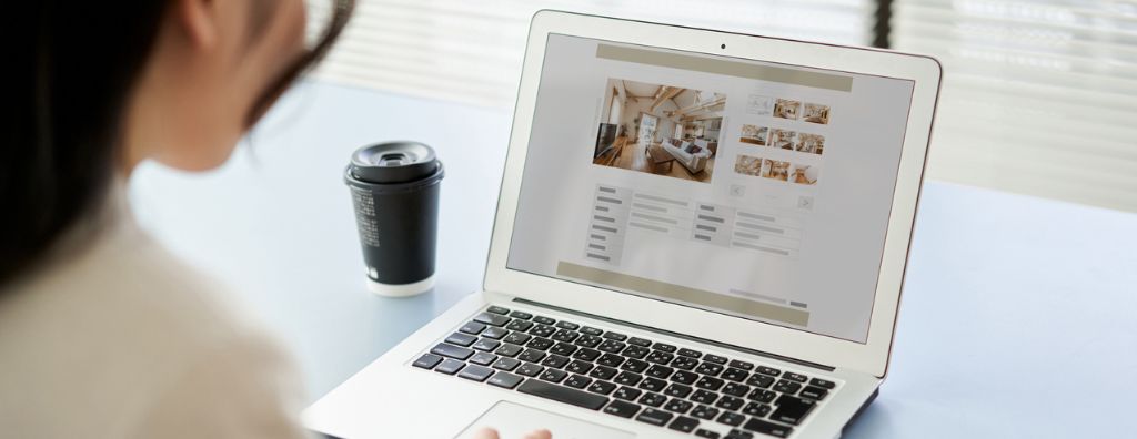 A young Asian woman first-time home buyer searches for homes online. She sits at her dining room table with a cup of coffee while she looks at real estate listings.