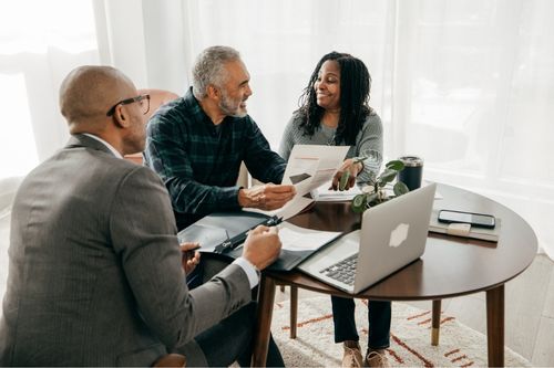 A Black heterosexual couple meet with their real estate agent to sell their home. They sit at a coffee table. The agent is a young Black man. He carries a notebook filled with paperwork, a laptop, and real estate paperwork.