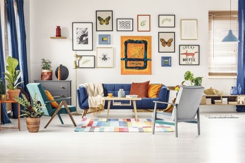 A living room decorated with eclectic interior design. It has a gallery wall above the couch with a variety of framed photography and hand drawings. The living room carpet is made up of dozens of pastel cubes, there are colorful pillows and houseplants throughout, and the furniture is minimalist.