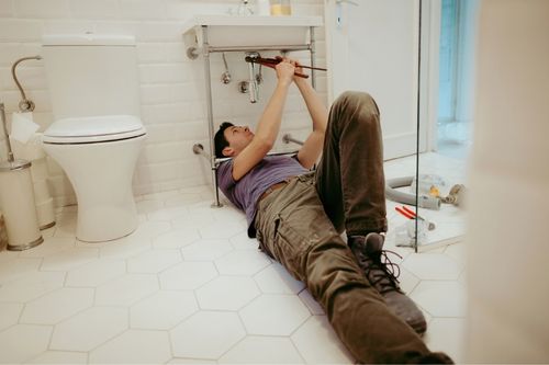 A young Caucasian man lays on his back in a bathroom with white hexagon tile flooring and white subway tile walls. A first-time home seller, he uses a wrench to turn the drainpipe under the bathroom sink as he prepares to sell his home.