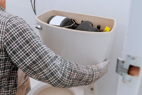 A closeup shot of a man in a plaid shirt preparing to fix a toilet in a bathroom with white tile walls. He has the tank open and is getting ready to remove the toilet from the floor.