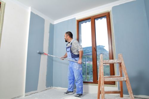 A middle-aged Caucasian man with dark hair paints bedroom walls sky blue. He wears painting overalls and uses a long-handled roller to apply the blue paint from the baseboard to the trim bordering the ceiling.