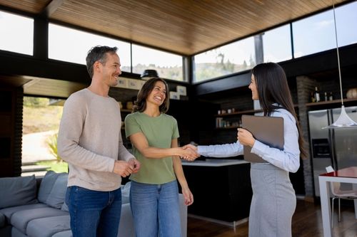 A young heterosexual Hispanic couple shake hands with their property manager in the open kitchen area of their modern investment home. The kitchen has black cabinets and stainless-steel appliances. The ceiling is hardwood and there is a band of windows around the unit just below it.