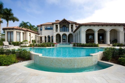 A mansion that incorporates elements of Greek Revival home design, with marble columns, terra cotta roofing, and large arched doorways. There is a large blue in the mansion’s front courtyard, with landscaping design woven throughout.