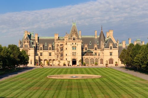 An aerial photo of The Biltmont Estate in Asheville, North Carolina. It is a chateau-style mansion built in the late 1800s. It is massive, with dozens of rooms visible from its front façade. The front lawn extends hundreds of feet out from the house, lined by trees on either side.
