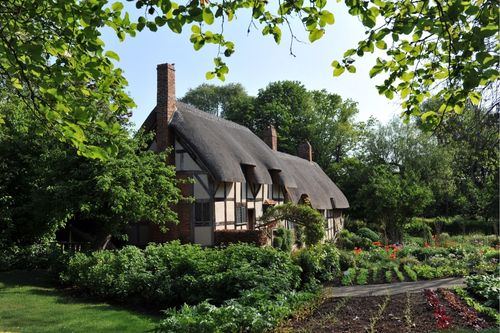 A profile shot of a brown and white English Tudor style house with a thatched roof and a tall chimney. The siding is made up of half-timbering wood patterns and brick accents. The garden surrounding the home is in full bloom, producing lettuce and spices.