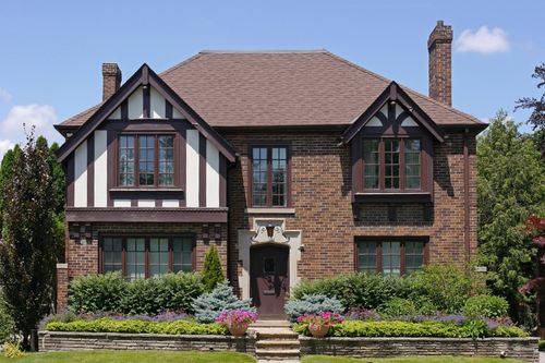 The front façade of a brown and white Tudor style house with interlocking gabled roofs, brick accents, a tall chimney, several windows, and a decorative front entrance with stone masonry framing the doorway. There are several shrubs and a hedge in the front yard garden beds.