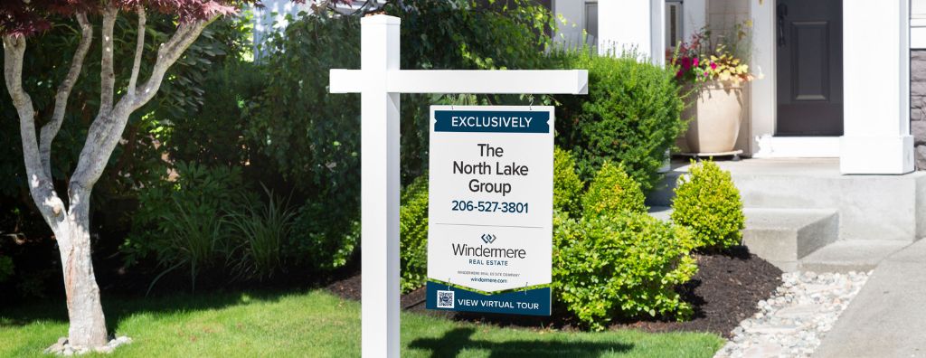 A Windermere Real Estate For Sale sign in front of a green shrub.