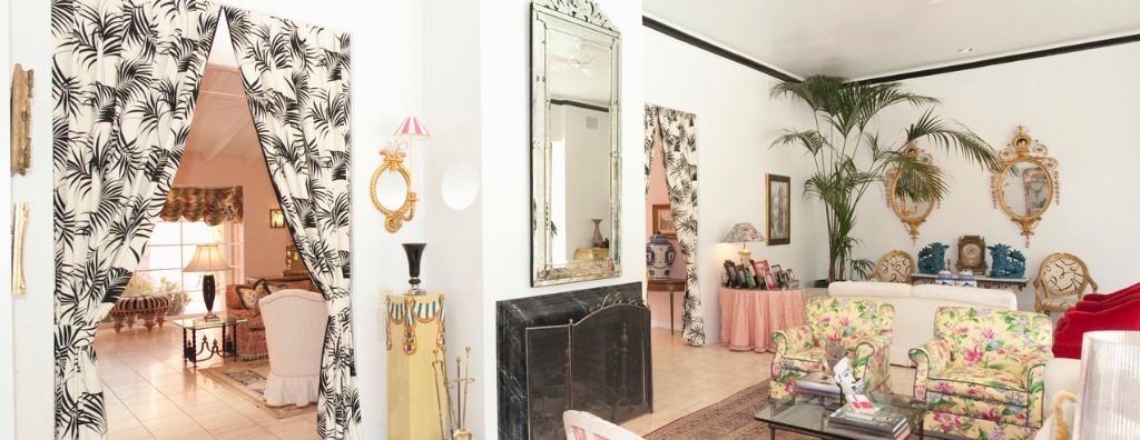 A living room decorated with eclectic interior design including palm tree pattern drapes hanging from the doorframes, floral print chairs, gold framed mirrors, and pink table covers.