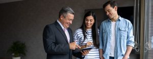 A heterosexual Hispanic couple smiles with their real estate agent looking at his tablet as they work together to sell their home. They are first-time home sellers. They all stand in a staged living room next to a window.