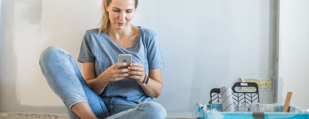 A young Caucasian woman with light hair sits on the floor of her living room as she prepares to paint the walls. She is one her phone, looking up how much paint to use.