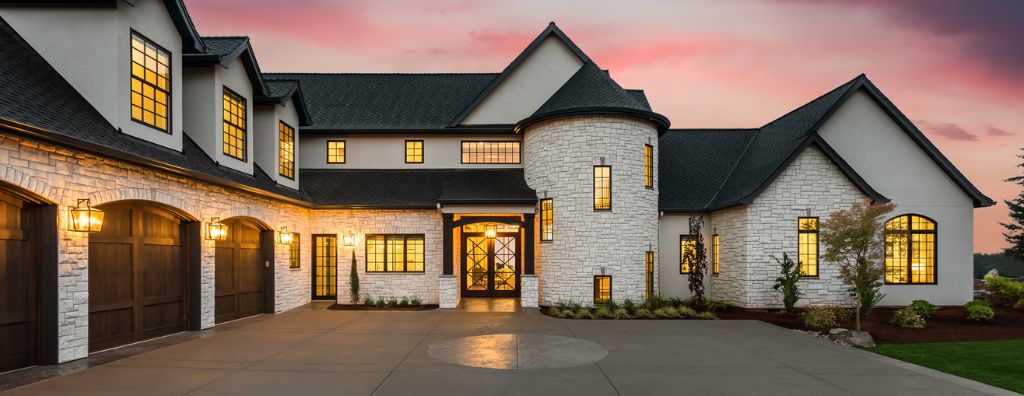 A white stone mansion in the countryside photographed at sunset with many warmly lit windows, a three-car garage, a wide driveway with embedded stonework, and a black rooftop. It has a medieval style, with parapet-type walls and a cylindrical room adjacent to the front door that looks like a castle tower.