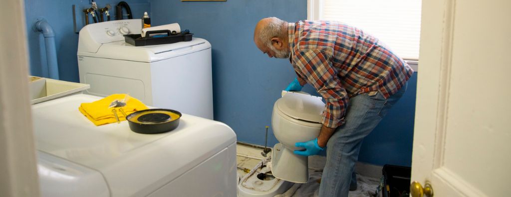 An older man fixes his toilet in his laundry room. He wears rubber gloves; his hand tools sit on top of the washer and dryer.