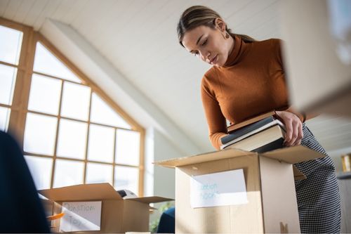 A young Caucasian woman starts to declutter before selling her home. She places books into a box labeled “donation books” in her home office.