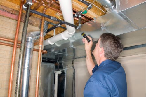 A Caucasian man home inspector works during the home buying process. He shines a flashlight at plumbing pipes in the basement.