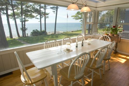 The dining room of a waterfront home, decorated with white traditional chairs and dinner table. The wall facing the lake is one large window with a view of the backyard and the lake just beyond it.
