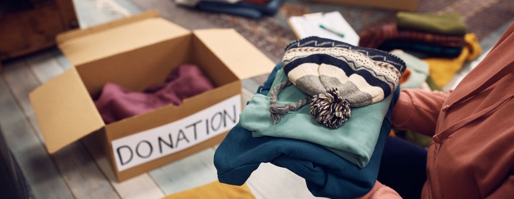 A woman starts to declutter before selling her home. She is organizing items in the living room. She holds a small pile of winter clothing and prepares to place it in a box labeled “donation” on the floor.