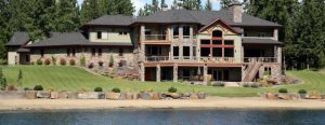 A luxury mountain lodge-style waterfront home with stone masonry, an A-frame window in the great room, and a wraparound deck. The backyard slopes down to the beachfront at the lake.
