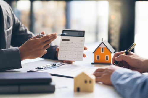 A closeup of two men’s hands doing paperwork at an office desk as they figure out how to pay for a house. One man points to a calculator while the other takes notes.