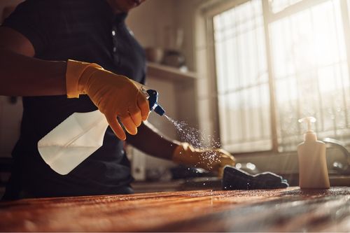 A closeup shot of a man’s arms cleaning his home. He sprays a bottle of cleaning solution on his kitchen countertop and scrubs the surface as the sunlight pours in through the window. He wears yellow rubber gloves and a navy polo shirt.