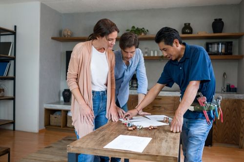 A Caucasian heterosexual couple are discussing a home remodeling project with a Latin American contractor as they prepare to sell their home. They look over paperwork on a wooden dining room table.