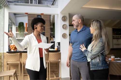 A Black woman real estate agent reviews a final walkthrough checklist with her clients, a Hispanic heterosexual couple, in the living room of a house.