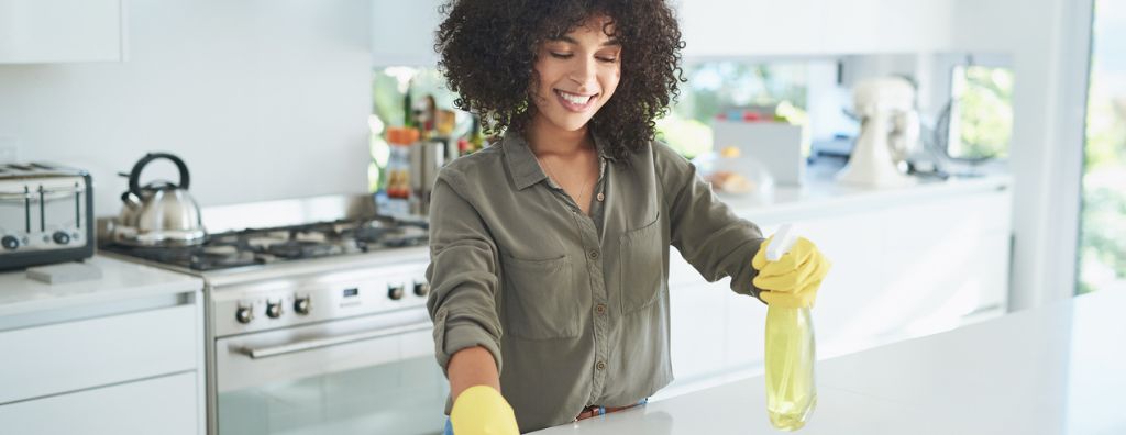 A young mixed-race woman wears yellow rubber gloves, spraying and wiping down her counter with natural cleaning products. Her kitchen has white marble countertops, stainless steel appliances, and white tile walls.