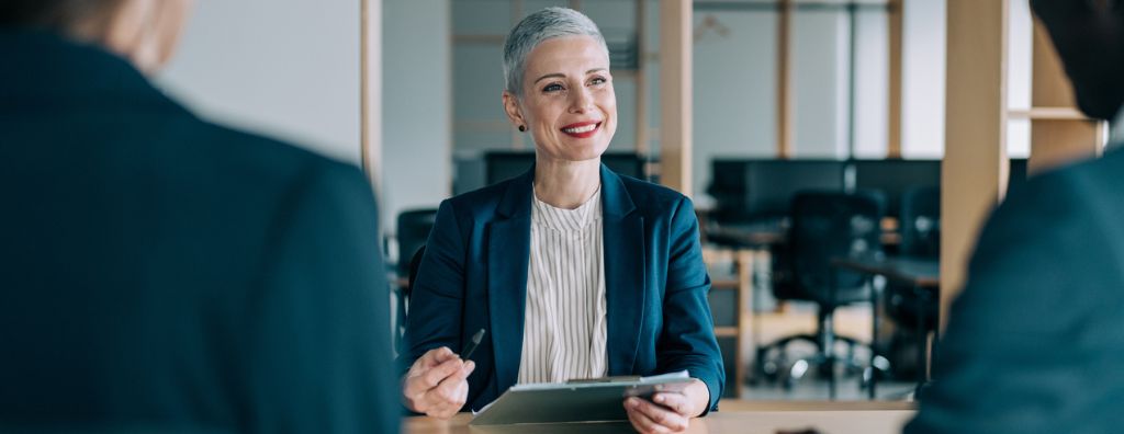 A White woman real estate agent with short white-grey hair sits at an office table with her two clients, who are blurred in the foreground so you can only see their shoulders. They prepare for the final walkthrough as they sell their home.