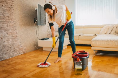 A young woman using home cleaning tips to eliminate common household odors. She scrubs her hardwood living room floor while listening to music.