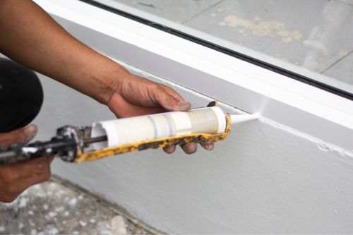 A closeup of someone’s hands doing home pest control prep work by caulking a new seal below a window.