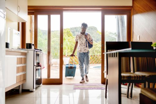 A young Black man walks into the living room of his second home, a modern loft with a large deck. He is rolling his suitcase behind him and holding a carry-on bag on his shoulder.