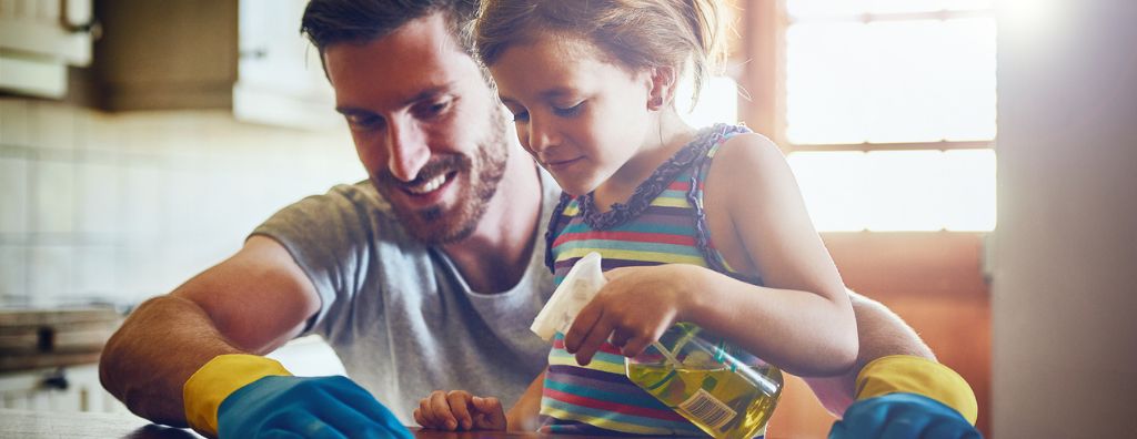 A father and his young daughter work together in the kitchen, using home cleaning tips to eliminate common household odors. The young daughter sprays a solution on the stovetop and the father scrubs with a sponge.