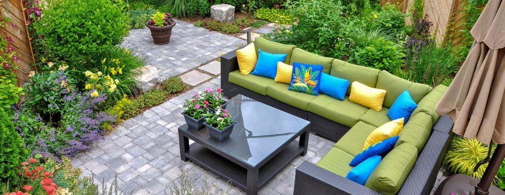 A backyard with stone patio pavers, a nice outdoor couch and slate coffee table, surrounded by lush flower beds with colorful plants. DIY projects.