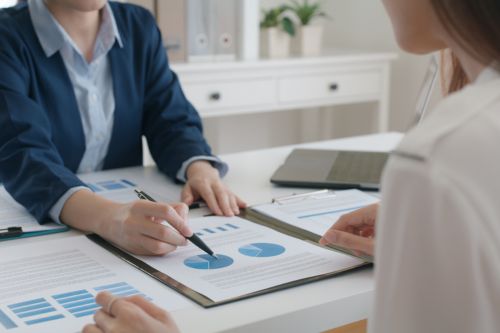 A real estate agent shows her female client how to negotiate as a seller. The agent is showing the clients charts of local real estate market conditions in a modern office.