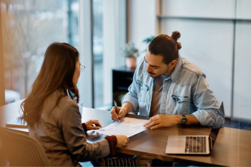 A young male home seller meets with his female real estate agent—one of the most important people in the home selling process—to discuss the sale of his home and sign paperwork.